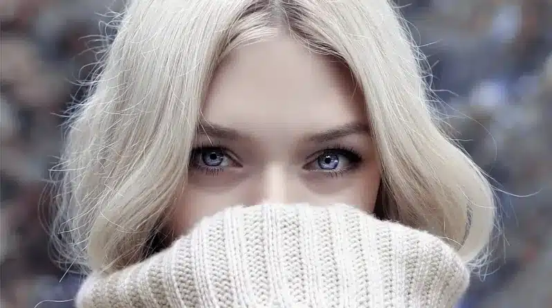 Nanobananaのプロンプトでポーズ変更する例。白いニットで口元を隠す、青い瞳が印象的な女性。